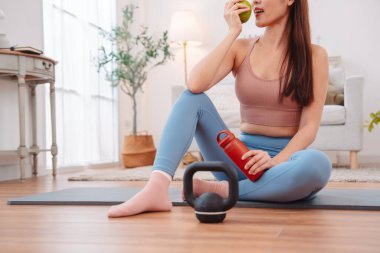 Kadın antremandan sonra mola veriyor, yoga minderinde halterler, kettlebell, yeşil elma ve su şişesiyle oturuyor. Sağlıklı yaşam tarzı, egzersiz sonrası iyileşme ve evdeki fitness beslenmesi..