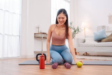 Kadın antremandan sonra mola veriyor, yoga minderinde halterler, kettlebell, yeşil elma ve su şişesiyle oturuyor. Sağlıklı yaşam tarzı, egzersiz sonrası iyileşme ve evdeki fitness beslenmesi..