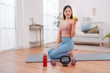 Kadın antremandan sonra mola veriyor, yoga minderinde halterler, kettlebell, yeşil elma ve su şişesiyle oturuyor. Sağlıklı yaşam tarzı, egzersiz sonrası iyileşme ve evdeki fitness beslenmesi..