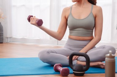 Genç Asyalı kadın evde dambıl ve kettlebell ile egzersiz yapıyor, yoga minderinde aktif giysiyle oturuyor. Sağlıklı yaşam tarzı, kapalı alanda çalışma, dayanıklılık eğitimi ve sağlık konsepti.