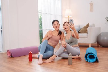 Asian Female friends relaxing together after workout at home, smiling and taking selfie with smartphone on yoga mats. Friendship, wellness lifestyle, rest, and social connection in a cozy setting.