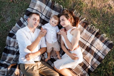 Anne, baba ve küçük oğlu yaz parkında battaniyenin üzerinde yatıyor. Yaz tatili kavramı. Anneler günü, babalar günü, bebekler günü. Aile doğada birlikte vakit geçiriyor. Aile görünüşü.