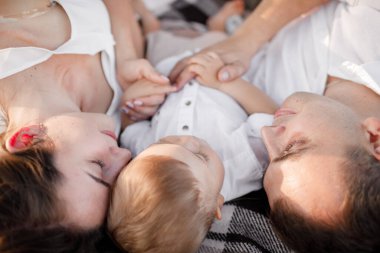 Anne, baba ve küçük oğlu yaz parkında battaniyenin üzerinde yatıyor. Yaz tatili kavramı. Anneler günü, babalar günü, bebekler günü. Aile doğada birlikte vakit geçiriyor. Aile görünüşü.