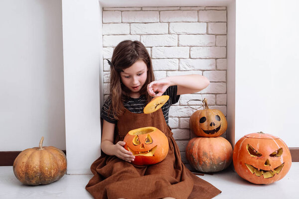 little girl with carving pumpkin on Halloween at home sitting next to fireplace in living room. Trick or treat. Child celebrating Halloween