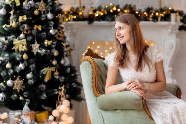 Güzel ve neşeli esmer kız açık bir gülümsemeyle Noel ağacının yanında güzel bir sandalyede oturuyor. Yeni yıl dekoru. Tatil kavramı.