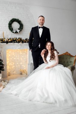 Sevgili gelin ve damat, Noel 'de Noel ağacıyla süslenmiş arka planda stüdyoda poz veriyorlar. Mutluluğun ve aşkın tadını çıkar.
