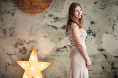 Enfes, çıplak ve dantelli bir elbise giymiş genç, sarışın bir kadın. Zarif elbiseli çekici bir kadın model. Tuğla duvara karşı poz veriyor. Arka planda büyük bir saat ve yıldız var.