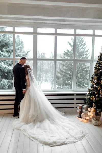 Young couple in love bride and groom posing in studio on background decorated with Christmas tree in their wedding day at Christmas near the large panoramic window. copy space