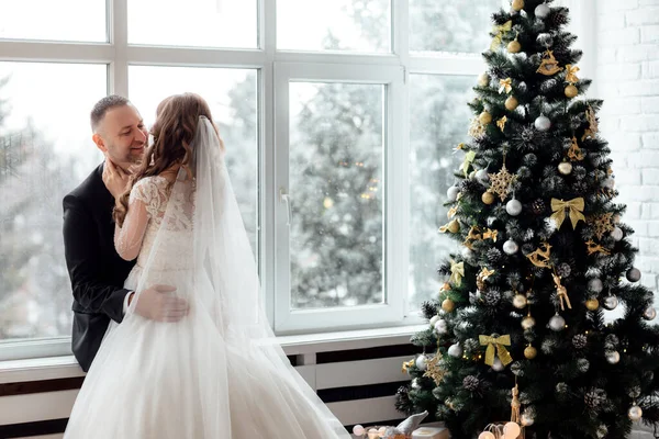 在他们的婚礼当天 一对年轻夫妇在大的全景窗前 在装饰着圣诞树的背景下 在摄影棚里摆着新郎和新娘的姿势 复制空间 — 图库照片