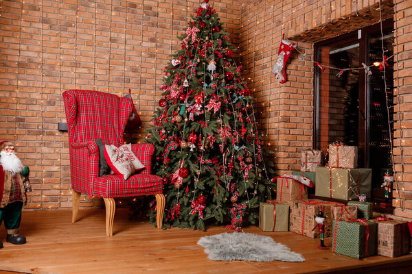 Room decorated to christmas celebration. christmas tree with red balls, bows, gift boxes near the window, cozy chair and santa near brick wall.