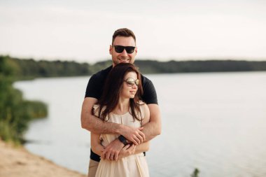 Göl kenarındaki sahilde mutlu aşıklar. Genç çift yaz günü gün batımında dışarıda sarılıyor. Birbirine aşık bir erkek ve kadın. Aşk ve aile kavramı. Tam boy. Kapat.