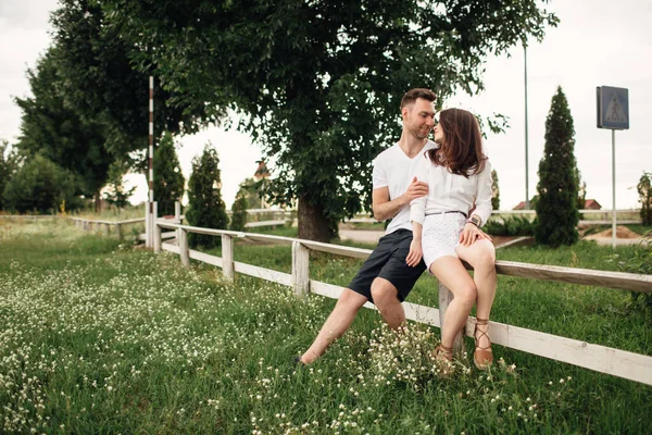 这对可爱的夫妇正坐在夏季公园的白色篱笆上 在温暖的日子里 男人和女人都在户外拥抱 — 图库照片