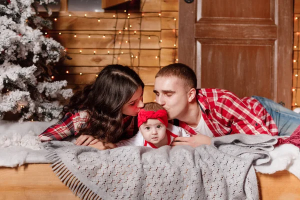 dad and mom kissing their baby daughter and lying on the bed near christmas tree. christmas time.