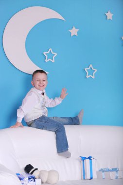 Noel tatili. Noel hediyesi kutusuyla beyaz bir kanepede oturan küçük bir çocuk. Arka planda beyaz ay olan mavi duvar