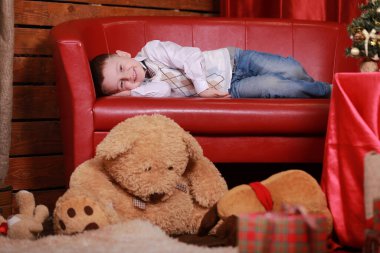 Küçük çocuk Noel stüdyosunun kırmızı koltuğunda eğleniyor. Arka planda Noel ağacı ve oyuncak ayı.