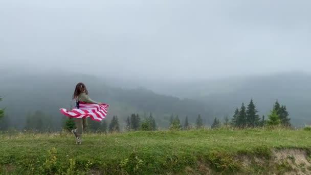 Gelukkig meisje rent en springt zorgeloos met open armen in troebele bergen. Ze houden de vlag van de VS vast. selectieve focus. — Stockvideo