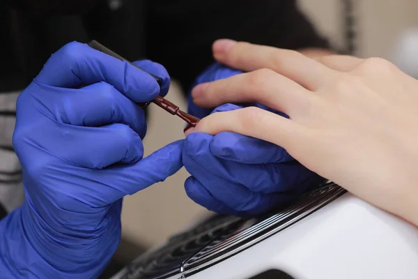 El maestro en los guantes protectores durante la manicura en el salón de belleza. La manicura maestra barniza el gel de marsala en las uñas de una clienta. El concepto de belleza y salud. —  Fotos de Stock