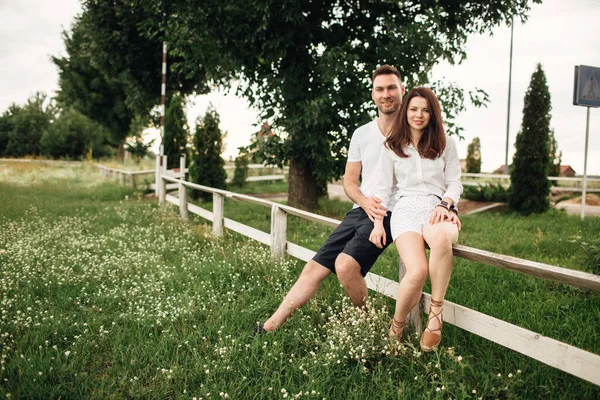 Sevgili çift yaz parkında beyaz çitlerde oturuyor. Erkek ve kadın sıcak bir günde dışarıda kucaklaşıyorlar..