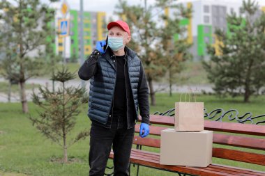 Kırmızı şapkalı, koruyucu maskeli ve paketli tıbbi lastik eldivenli teslimatçı müşteriye göndermek için akıllı telefon kullanıyor. Coronavirus sırasında teslimat servisi. İnternetten alışveriş.