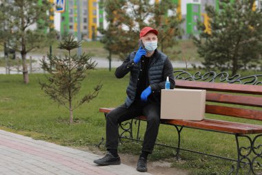 Kırmızı şapkalı, koruyucu maskeli ve paketli tıbbi lastik eldivenli teslimatçı müşteriye göndermek için akıllı telefon kullanıyor. Coronavirus sırasında teslimat servisi. İnternetten alışveriş.