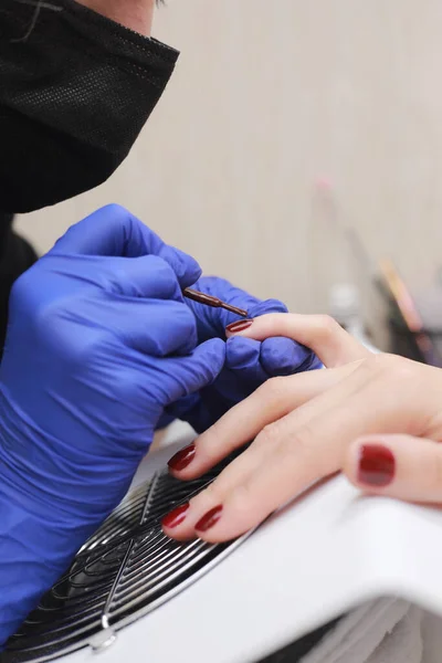 Master in protective gloves during a manicure at beauty salon. Master manicurist varnishes the marsala gel on the nails of a female client. The concept of beauty and health