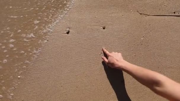 Kadın ıslak bir kuma kalp çizer. Dişi eller kumun üzerine deniz kelimesini yazıyor. Deniz suyu, kumlu bir sahildeki çizimleri temizler.. — Stok video