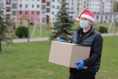 Koruyucu maskeli teslimatçı, eldivenler ve Noel Baba 'nın şapka kutusu açık havada, Coronavirus zamanında teslimat servisi.