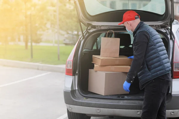 Young delivery man in protective mask, red cap and gloves near the car with boxes and packages, outdoors. Service coronavirus. Online shopping. mock up