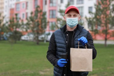 Kırmızı şapkalı teslimatçı, yüzü tıbbi maskeli, eldivenli kağıt torbaları tutan ve şehir dışında tek kullanımlık bardakta içen. Hizmet koronavirüsü. İnternetten alışveriş.