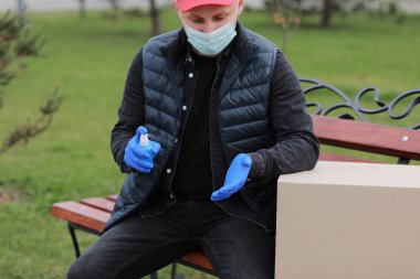 Coronavirus sırasında teslimat servisi kuryesi, COVID-19, salgın, tıbbi koruyucu maskeli teslimatçı, dışarıda karton kutuların yanındaki ellere alkol dezenfektan spreyi sıkan eldivenler..