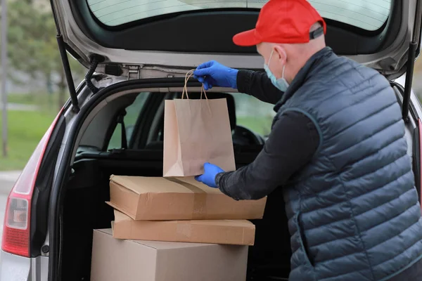 Delivery man in red cap, face medical mask and gloves hold paper bag near the car outdoor. Service coronavirus. Online shopping. mock up