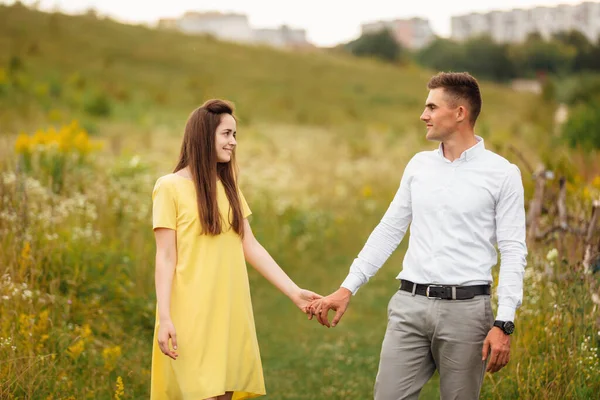 晴れた日に草の畑で手をつないでいる若い幸せなカップル 黄色のドレスの女の子と髪型の男 田舎を歩く夫婦の肖像画です — ストック写真