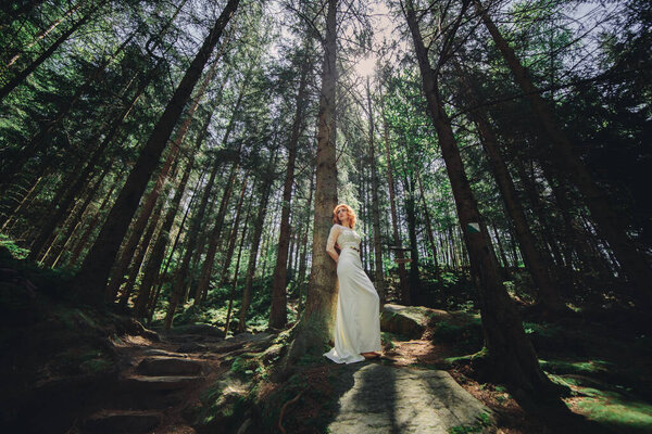 Young red hair bride in white long dress in a green forest on summer day