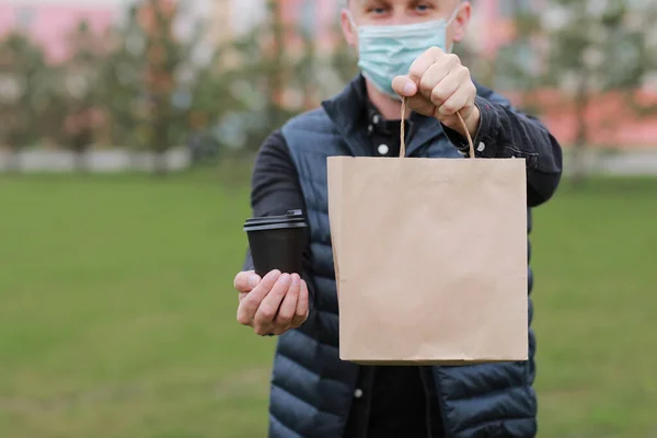 Delivery man in red cap, face medical mask hold take away paper bag and drink in disposable cup outdoors in city. Service coronavirus. Online shopping.