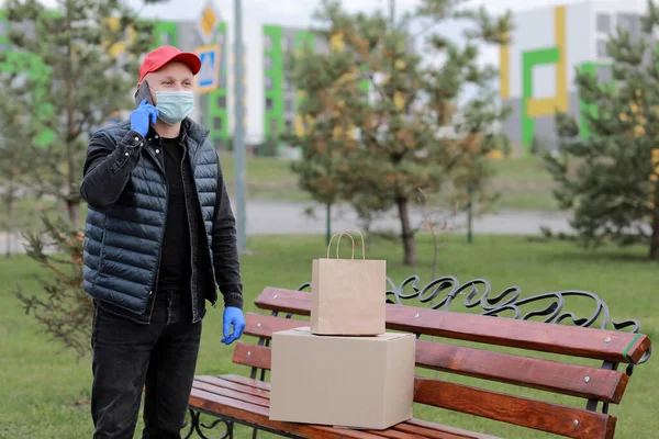 Kırmızı şapkalı, koruyucu maskeli ve paketli tıbbi lastik eldivenli teslimatçı müşteriye göndermek için akıllı telefon kullanıyor. Coronavirus sırasında teslimat servisi. İnternetten alışveriş.