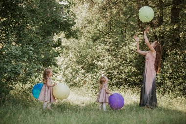 İki kızı olan güzel mutlu anne yeşil parkta eğleniyor ve yaz güneşli bir günde renkli balonlar tutuyor. Mutlu bir aile. Anne, çocuk, anne
