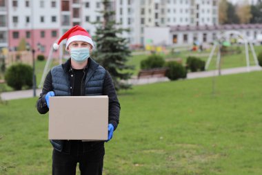 Koruyucu maskeli teslimatçı, eldivenler ve Noel Baba 'nın şapka kutusu açık havada, Coronavirus zamanında teslimat servisi.