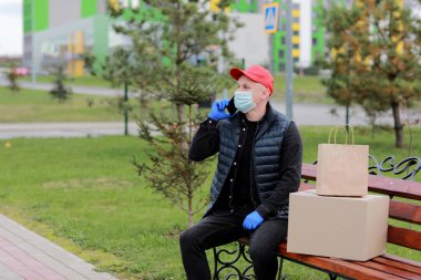 Kırmızı şapkalı, koruyucu maskeli ve paketli tıbbi lastik eldivenli teslimatçı müşteriye göndermek için akıllı telefon kullanıyor. Coronavirus sırasında teslimat servisi. İnternetten alışveriş.