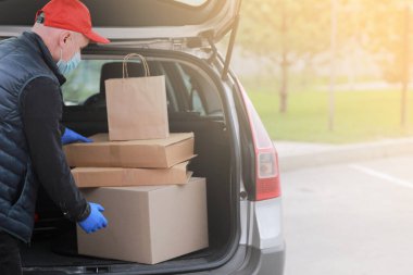 Koruyucu maskeli, kırmızı şapkalı ve eldivenli genç teslimatçı dışarıda kutular ve paketlerle arabanın yanında. Hizmet koronavirüsü. İnternetten alışveriş. maket yap