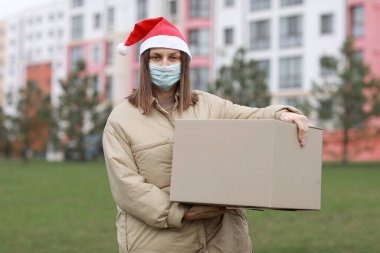 Kırmızı Noel Baba şapkalı ve tıbbi koruyucu maskeli teslimatçı kız dışarıda büyük bir kutu tutuyor. Yılbaşı kutusunda yeni yıl paketi, karantina zamanında online dağıtım mağazası. Hizmet koronavirüsü