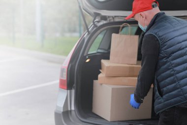Koruyucu maskeli, kırmızı şapkalı ve eldivenli genç teslimatçı dışarıda kutular ve paketlerle arabanın yanında. Hizmet koronavirüsü. İnternetten alışveriş. maket yap