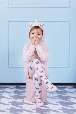 Tek boynuzlu at kostümlü güzel küçük bir kız gülümsüyor ve mavi duvarda eğleniyor. Bebek pembe büyük kutuda oturuyor. Mutlu çocukluk