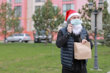 Noel üniformalı teslimatçı, koruma maskesi takan teslimat kağıdı taşıyan akıllı telefonu müşteriye gönderen adam. 2021 'de karantina sırasında yiyecekleri kaldırın.