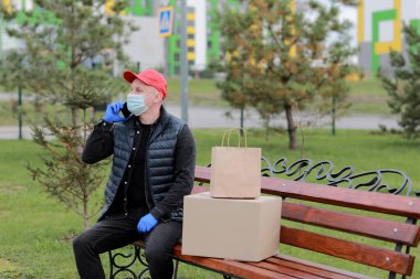 Kırmızı şapkalı, koruyucu maskeli ve paketli tıbbi lastik eldivenli teslimatçı müşteriye göndermek için akıllı telefon kullanıyor. Coronavirus sırasında teslimat servisi. İnternetten alışveriş.