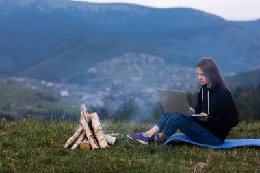 Genç bayan serbest yazar akşamları dağlarda dizüstü bilgisayarda çalışıyor. Kamp ateşinin yanında oturan turist kız eğleniyor. Boşluğu kopyala