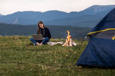 Genç bayan serbest yazar akşamları dağlarda dizüstü bilgisayarda çalışıyor. Kamp ateşi ve çadırın yanında oturan turist kız. Boşluğu kopyala.