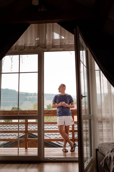 Ung man står på balkongen på hotellet på morgonen. hålla en kopp kaffe eller te i hennes händer. Utanför naturskog och berg — Stockfoto
