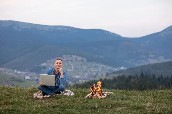 Dışarıda dizüstü bilgisayarla çalışan bir adam dağlarda oturuyor. Uzaktan çalışma ya da serbest yaşam tarzı kavramı. Hücresel ağ geniş bant kapsama alanı. İnternet 5G. Yürüyüşçü turist vadi manzarasının tadını çıkarıyor