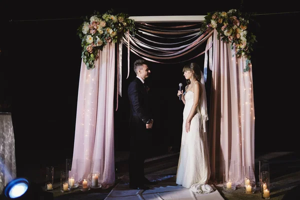 时尚的新婚夫妇晚上站在背光下 背对着别致的装饰和花朵 电灯泡和花环 入夜仪式植物学 薄膜效果 — 图库照片