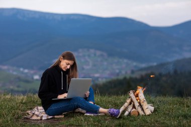 Genç bayan serbest yazar akşamları dağlarda dizüstü bilgisayarda çalışıyor. Kamp ateşinin yanında oturan turist kız eğleniyor. Boşluğu kopyala